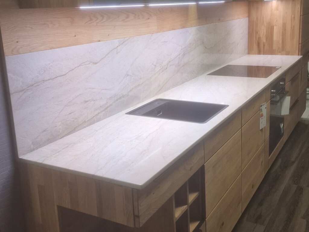 Holzküchen in Dresden: Massivholz, Furnier oder Holzoptik 22 Seitenansicht der Decker-Küche mit heller Natursteinplatte, Unterbauspüle und Rückwand aus demselben Stein, Licht unter den Oberschränken, Eichenfronten und offenen Fächern am Ende der Zeile